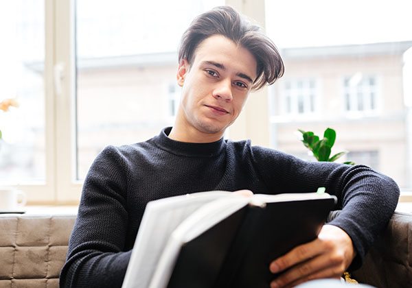 Image of cheerful caucasian student in library learning education material with books. Look at camera.